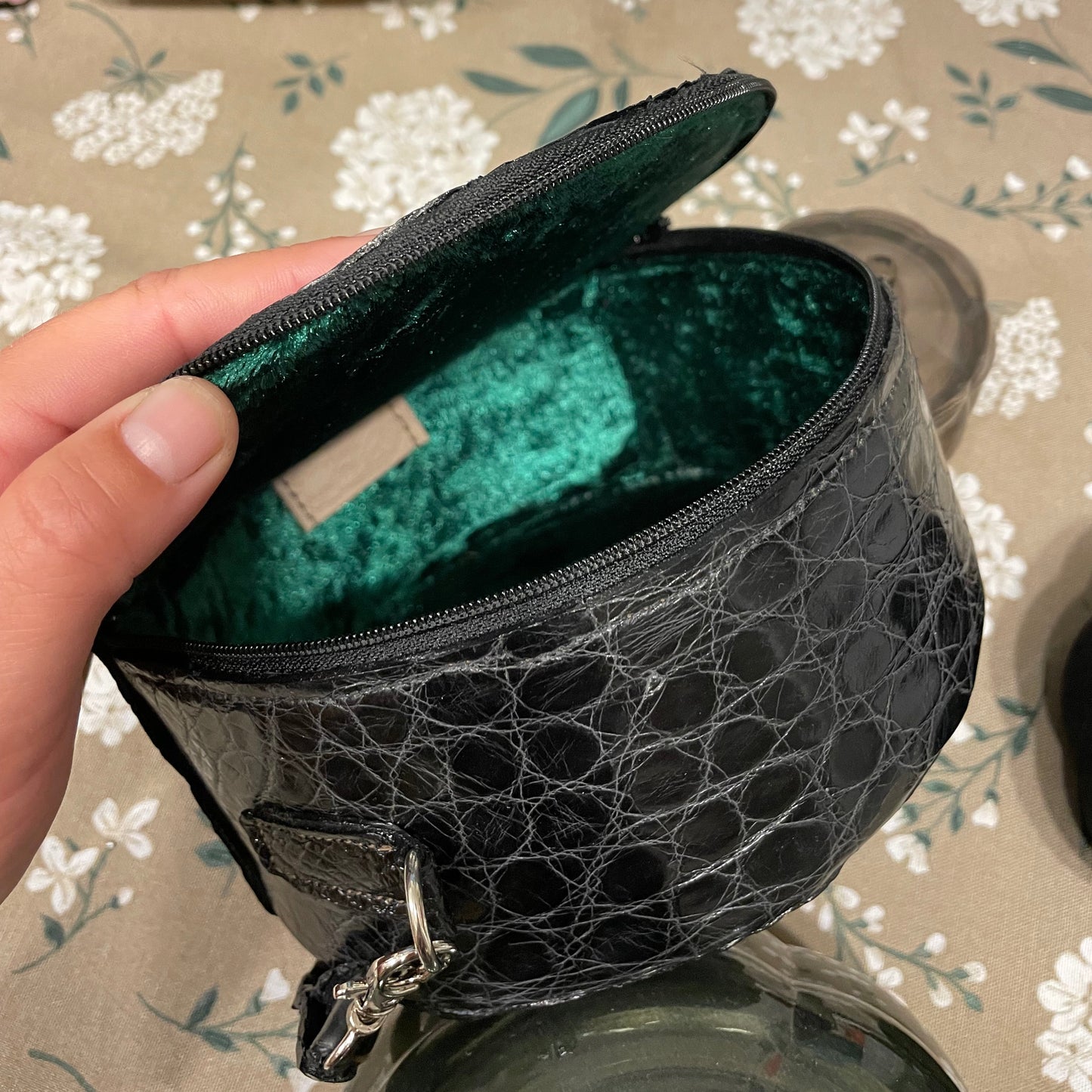 Black alligator skin purse with green interior held by a hand on a floral-patterned tablecloth.